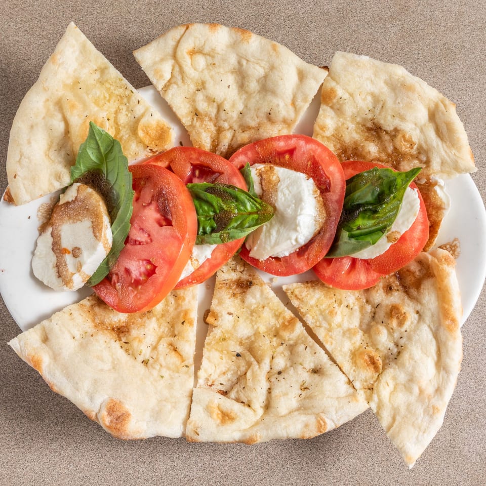 Caprese salad with sliced tomatoes, mozzarella, basil, balsamic glaze, and flatbread on a white plate.
